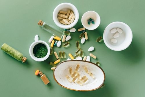 Vitaminas y suplementos en cápsulas esparcidos sobre fondo verde con utensilios.